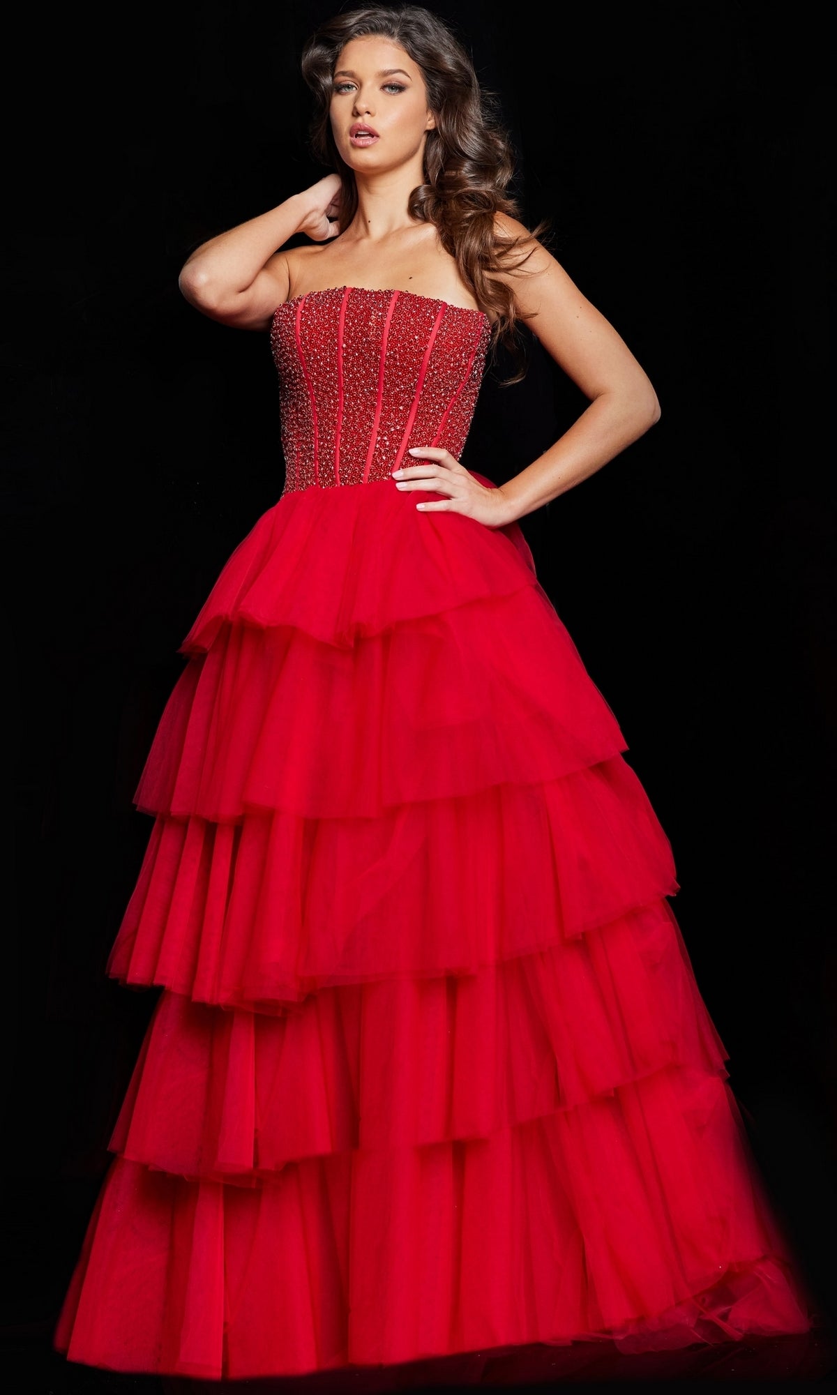 A woman wears the Long Prom Dress 37210 by Jovani, featuring a strapless tulle ball gown design with an embellished corset bodice and layered skirt, standing against a black background.